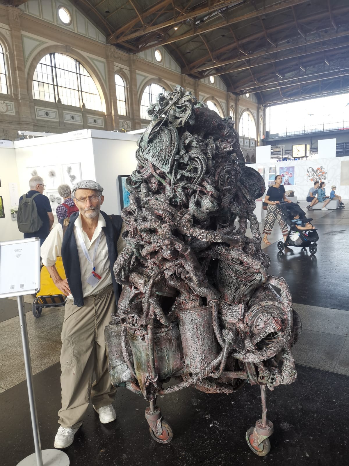 Hohe braun-graue Metallskulptur auf Rollen in einer Messehalle mit Bogenfenstern