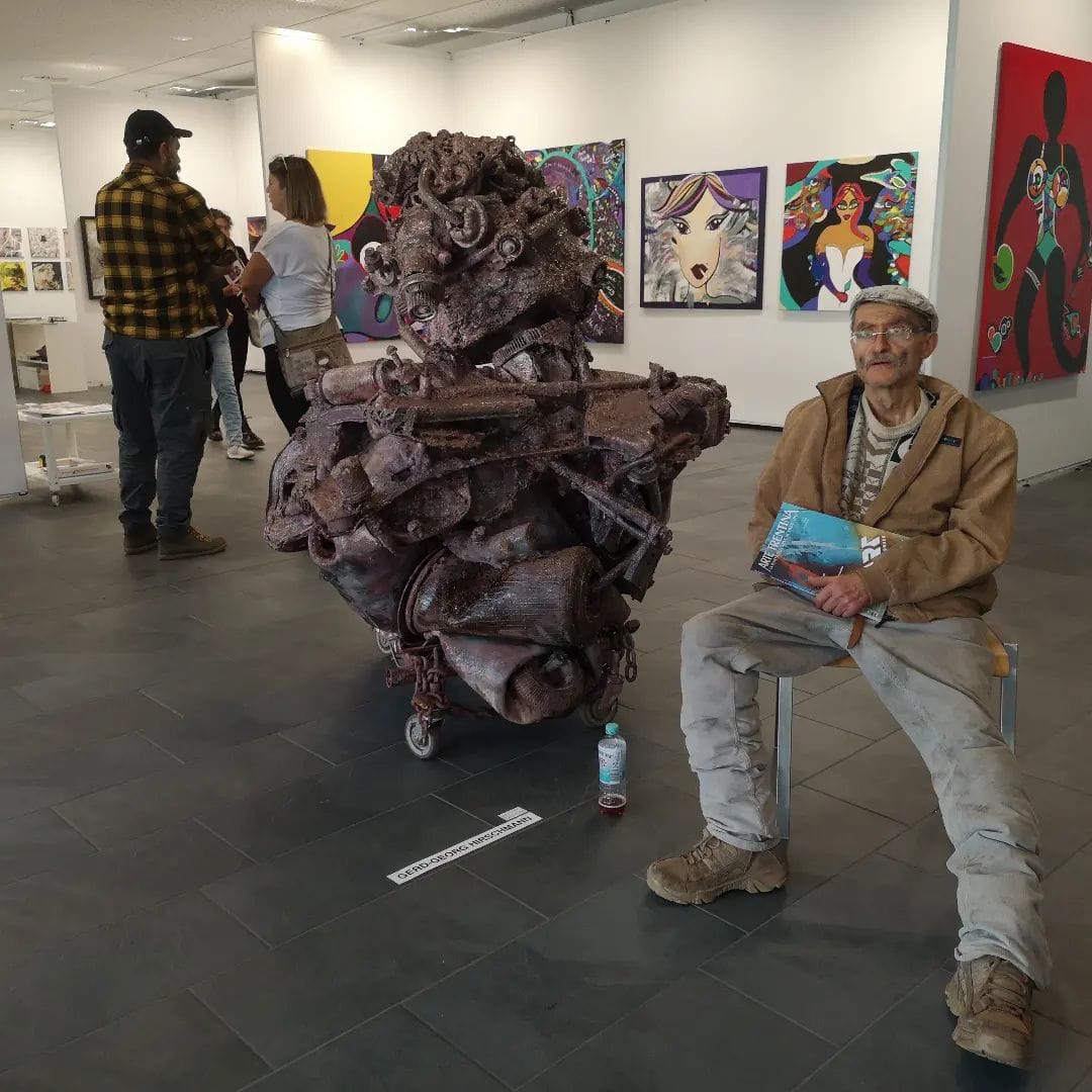 Braune Metallskulptur auf Rollen in einer weissen Galerie, dahinter Bilder an der Wand