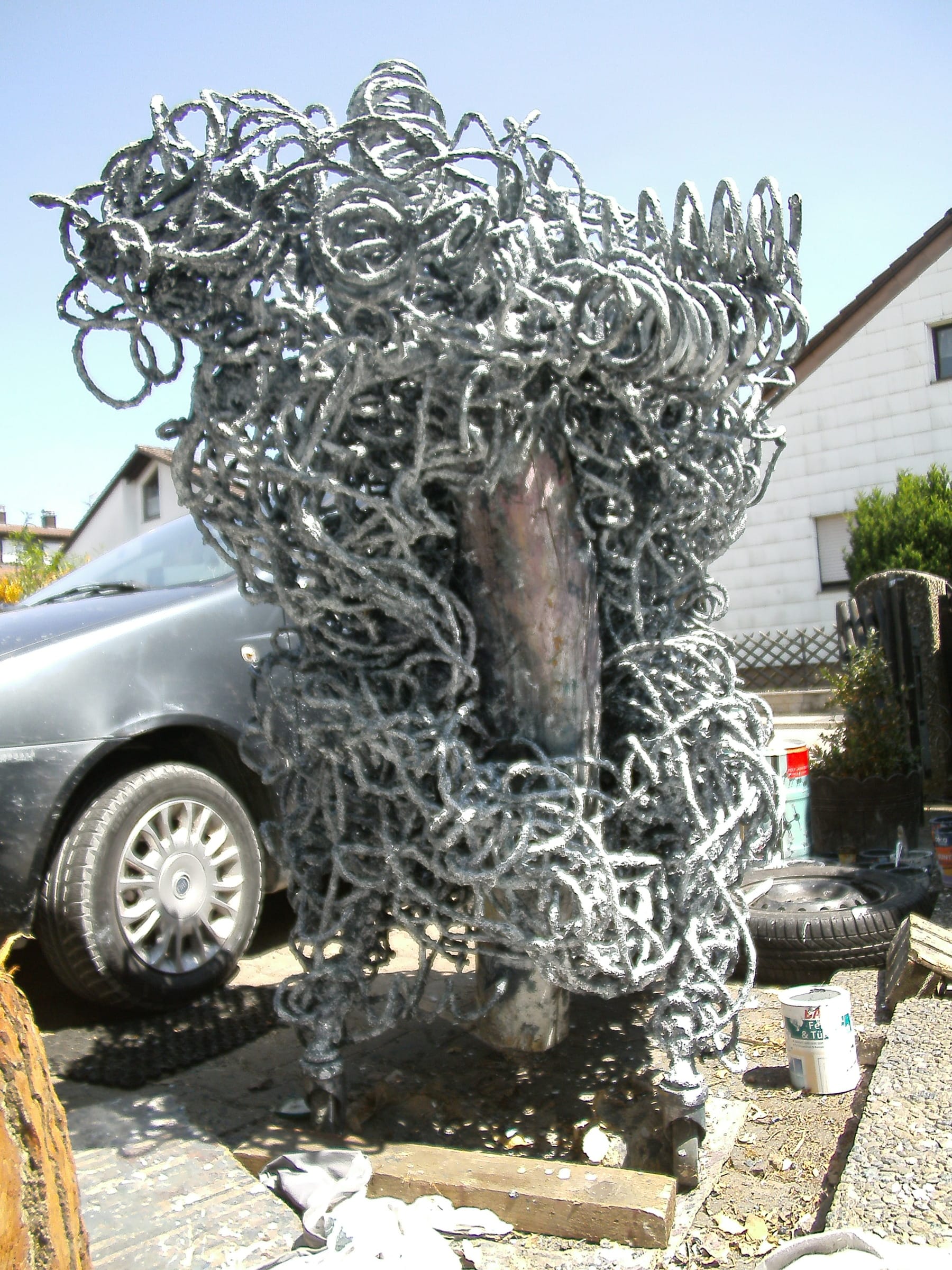 Hohe silbergraue Schlingen-Skulptur mit kupferfarbenem Kern in der Mitte, im Hof vor einem Haus und einem Auto