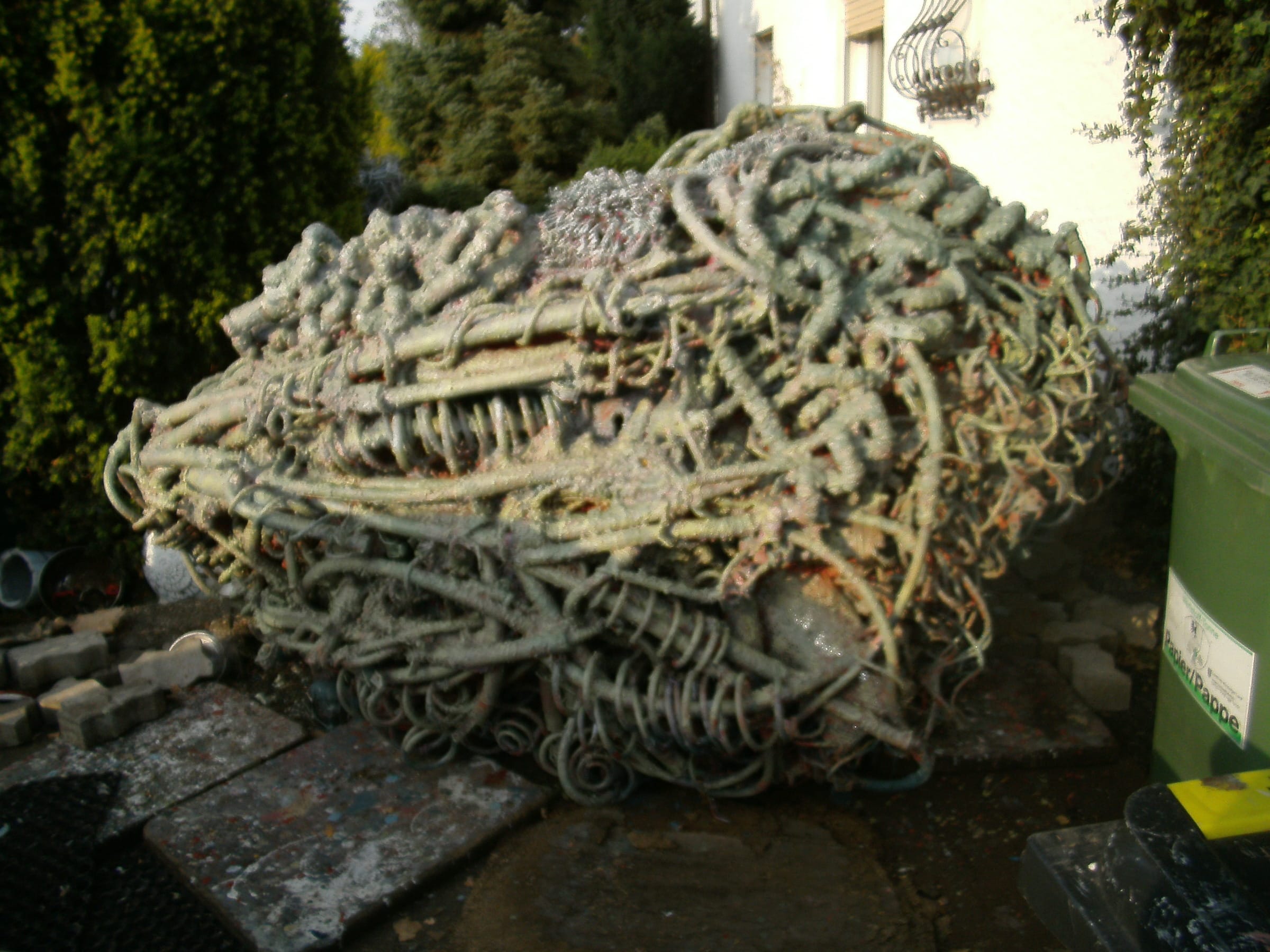 Liegende grün-graue Skulptur aus Metallschlingen im Hof vor einer Hecke