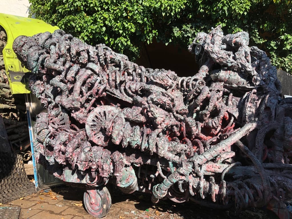Lange, wagenartige Skulptur auf Rollen, violett-rot-grün patiniert, voller Zahnräder und Metallteile, vor einer Hecke