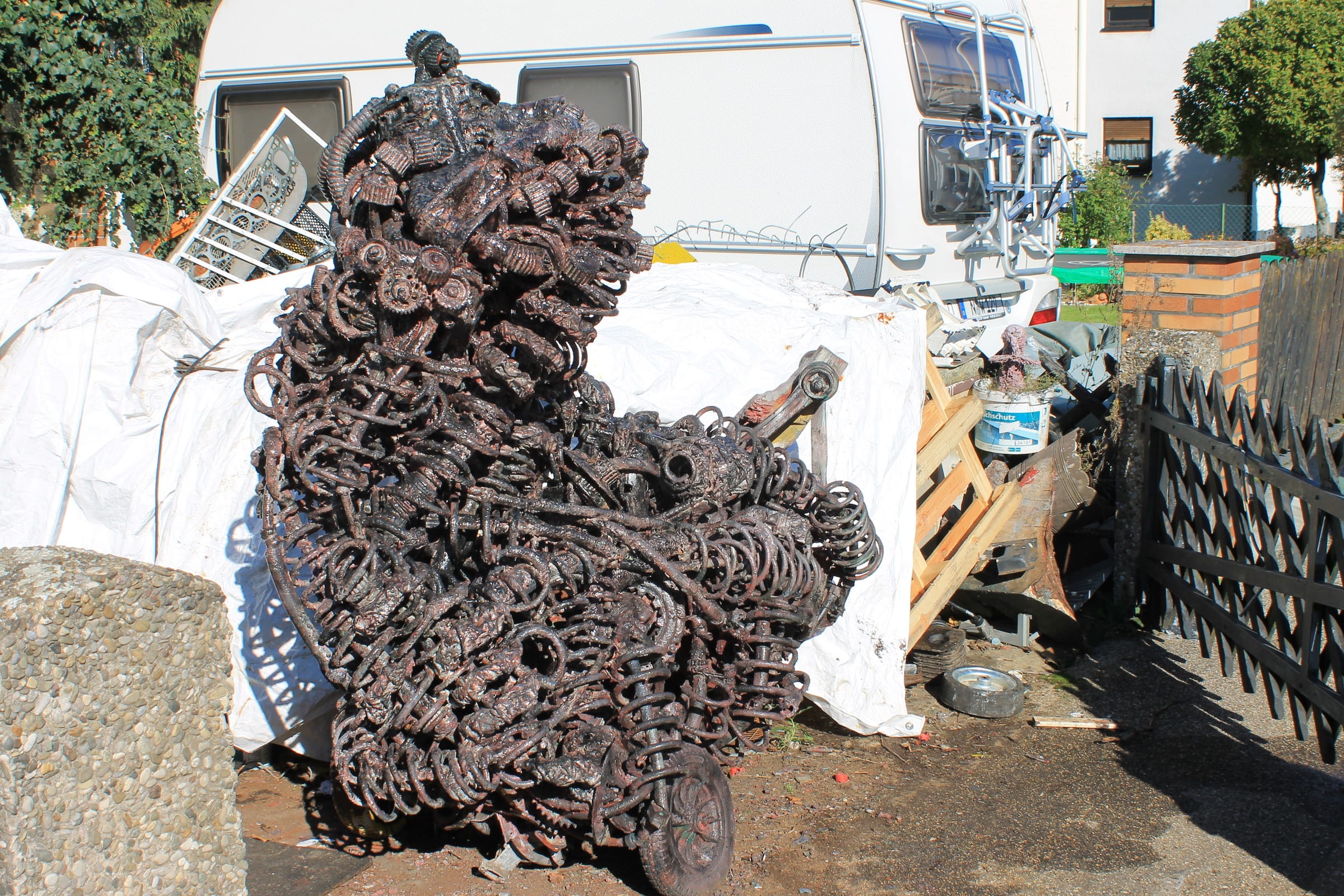 Kupferrote Metallskulptur aus Federn, Zahnrädern und Metallteilen auf Rollen, im Hof vor weissen Planen und einem Wohnwagen