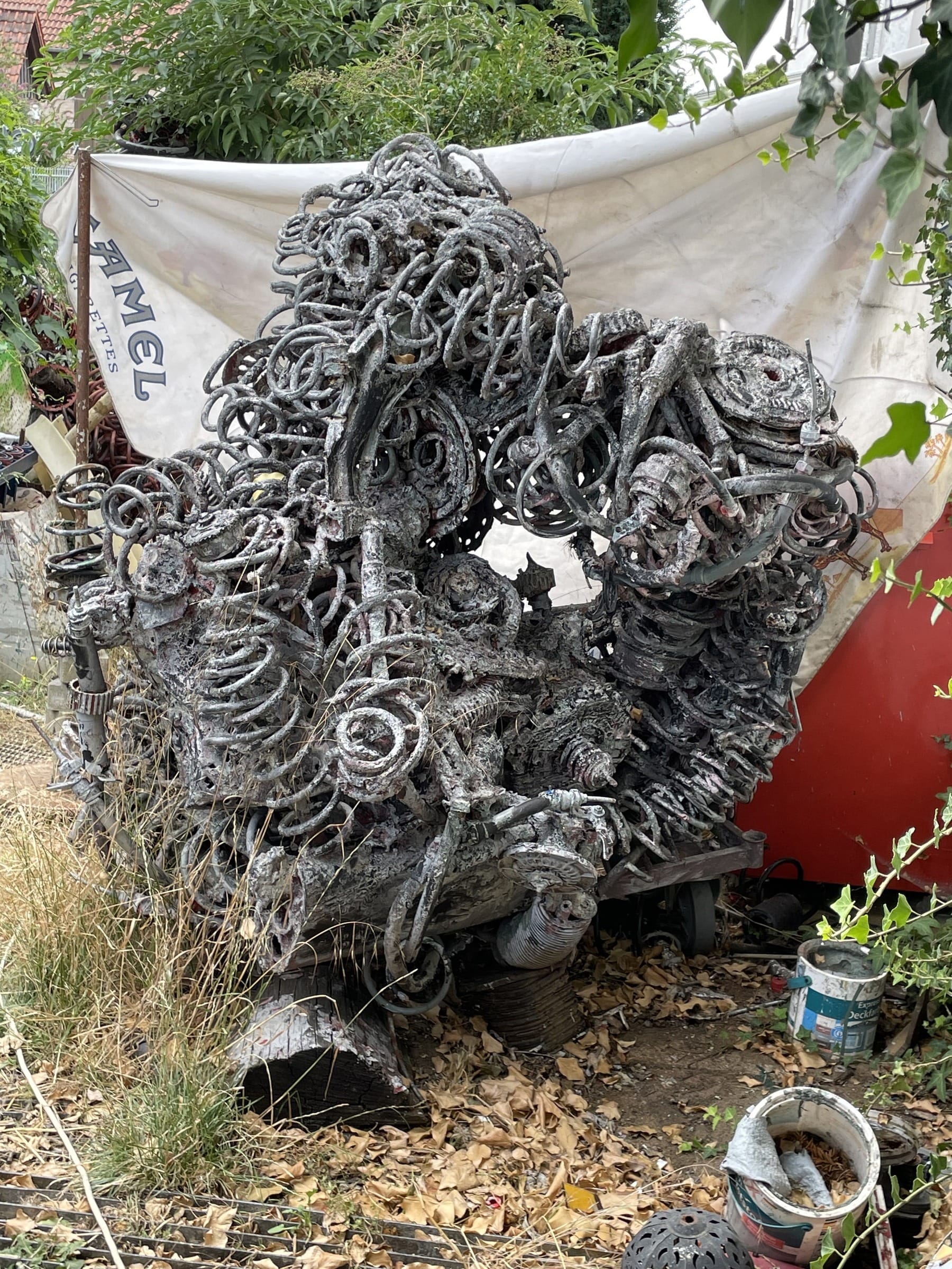 Silbergraue Skulptur aus Federn und Spiralen in einem verwilderten Garten vor einer weissen Plane