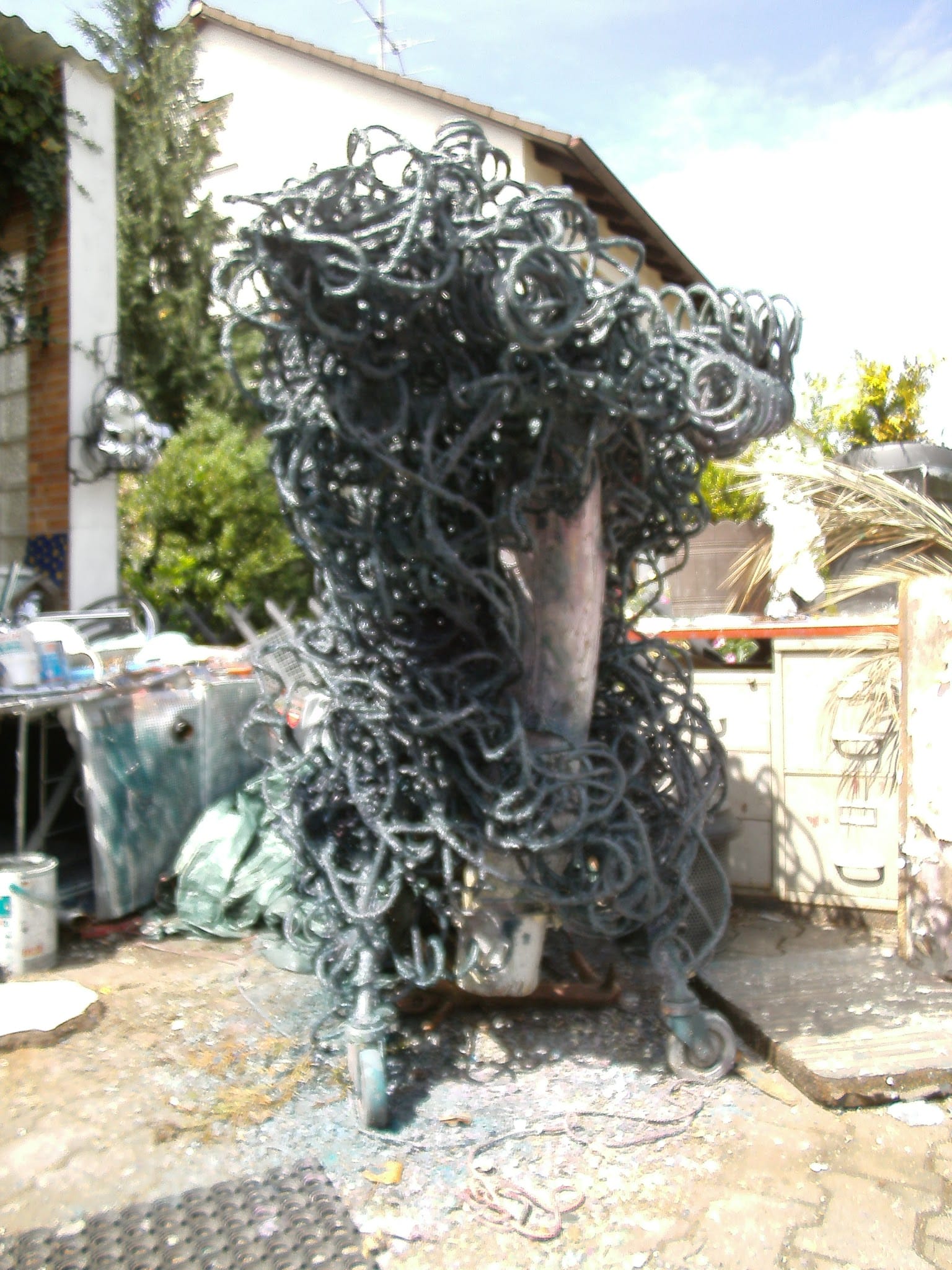 Hohe dunkel-grüngraue Skulptur aus Metallschlingen auf Rollen im Hof