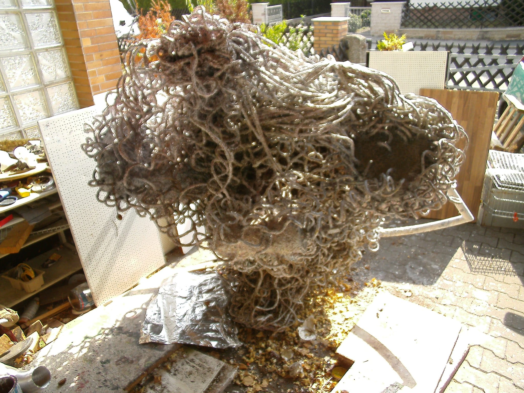 Hell-silbrige Skulptur aus Metallschlingen im Hof vor einer Wand aus Glasbausteinen