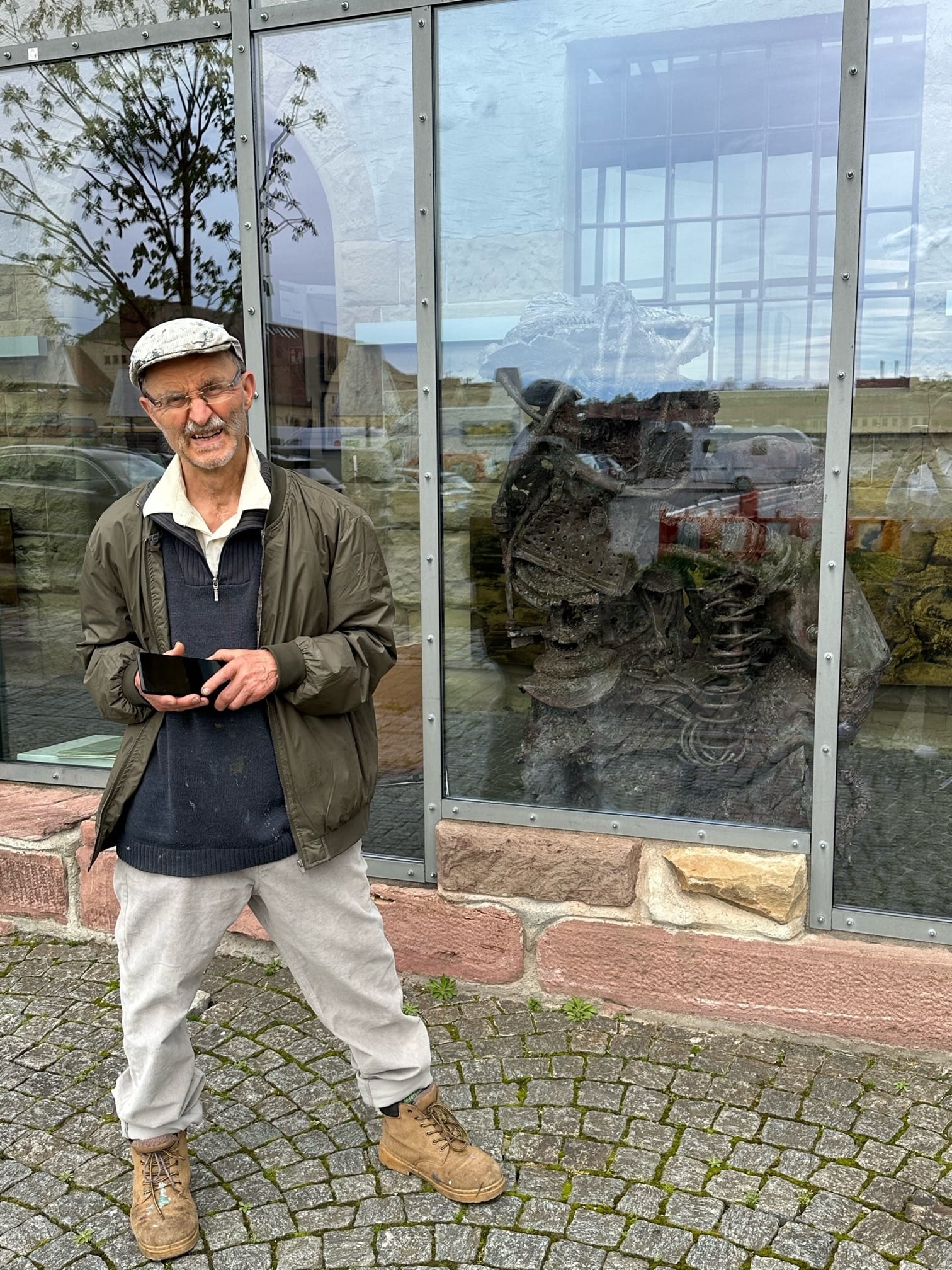 Gerd Georg Hirschmann mit Schiebermütze und grüner Jacke auf Kopfsteinpflaster vor einer Glasfassade, hinter der Scheibe steht eine Metallskulptur