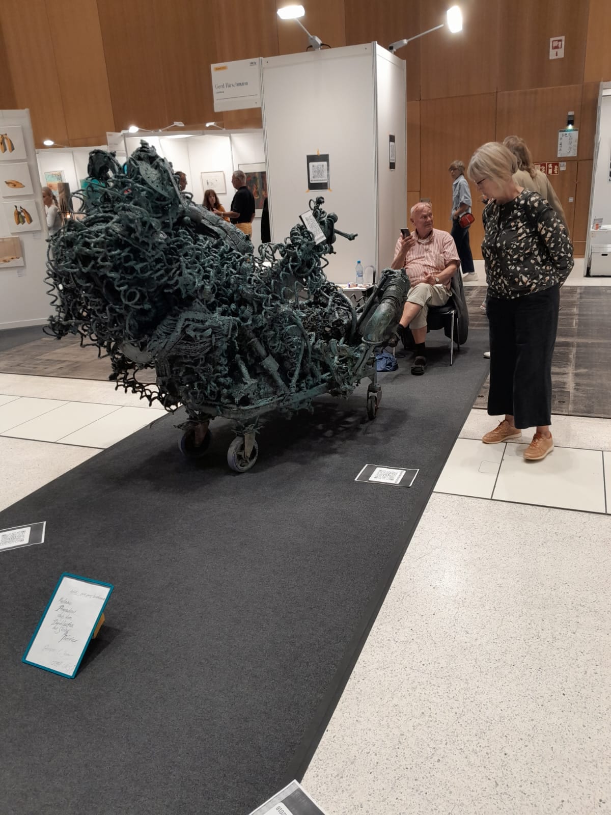 Grosse grün patinierte Metallskulptur auf Rollen an seinem Stand auf der ARTe Wiesbaden, Besucher davor